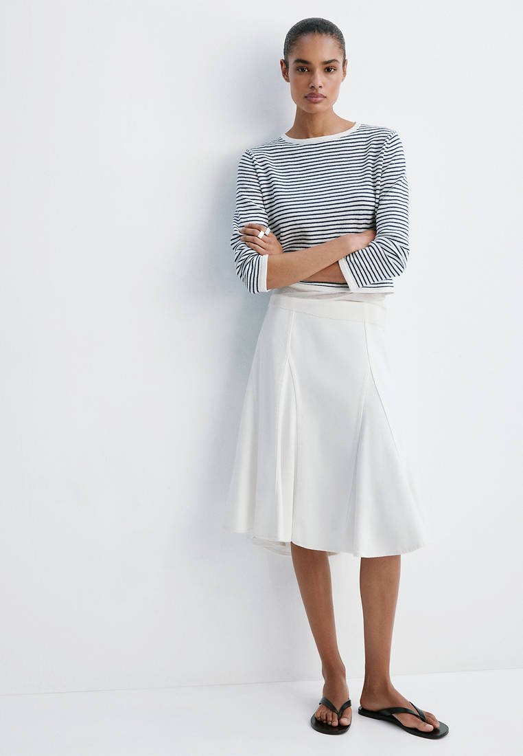 Femme debout, bras croisés, portant un haut à manches longues rayé noir et blanc, une jupe midi blanche et des sandales noires.