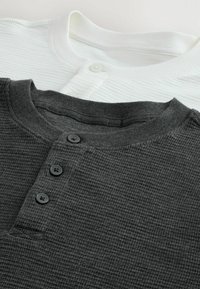 Due magliette Henley impilate; una nera con tessuto testurizzato e tre bottoni scuri, l'altra bianca con superficie liscia e un solo bottone.