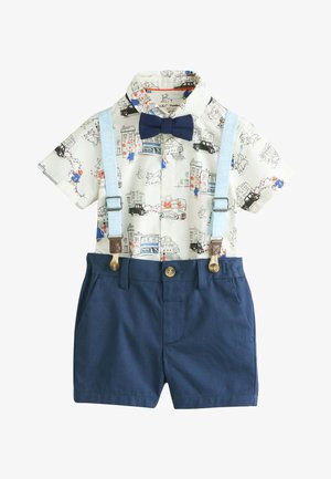 Bambino piccolo con capelli ricci che indossa una camicia a fantasia, papillon nero, bretelle azzurre e pantaloni scuri, in piedi in casa.