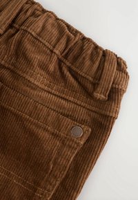 Pantalon en velours côtelé marron avec taille élastique, poche avant, passants pour ceinture et détail bouton en métal sur fond blanc.