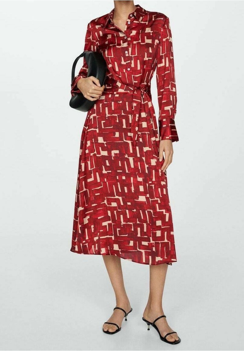 Femme portant une robe midi rouge à motifs avec manches longues, tenant une pochette noire et chaussée de talons noirs à brides sur un fond uni.