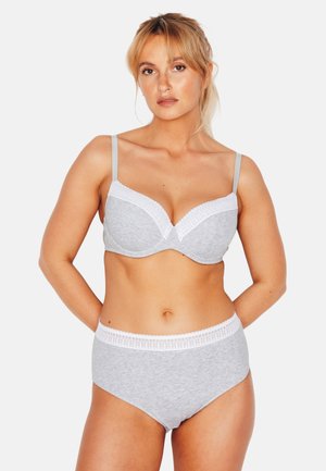 Femme aux cheveux blonds portant un soutien-gorge gris clair et une culotte taille haute avec bordure en dentelle blanche, debout devant un fond uni.