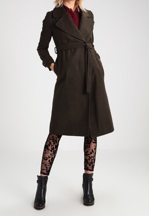 Kobieta w ciemnobrązowym płaszczu z paskiem, na bordowej bluzce, przezroczystych czarnych legginsach z wzorem i czarnych botkach do kostki, stojąca z rękami w kieszeniach.