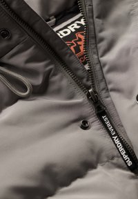 Grå pufferjakke med lynlås og knaplukning, struktureret stof og "Superdry Everest"-mærkat. Indvendig synlig med logofarver.