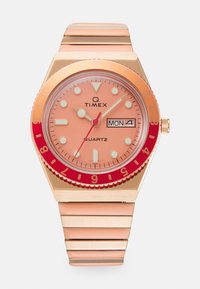 Montre Timex Q avec un cadran pêche, une lunette rouge, des accents argentés, un affichage jour/date et un bracelet en métal rose comportant des maillons segmentés.
