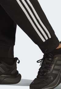 Čierne športové tepláky s bielym trojpruhovým dizajnom na boku, v kombinácii s čiernymi športovými topánkami s textúrovanými detailmi a šnúrkami.