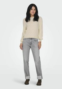 Suéter de punto beige claro con patrón de diamantes, combinado con jeans gris claro remangados y botas de tobillo marrones. Fondo plano.