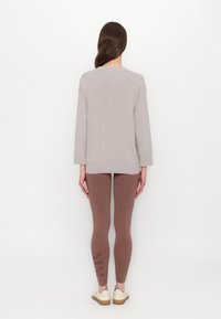 Lys grå ribbestrikket genser med avslappet passform, kombinert med brune leggings med mønster på siden. Modellen har på seg lyse sko.