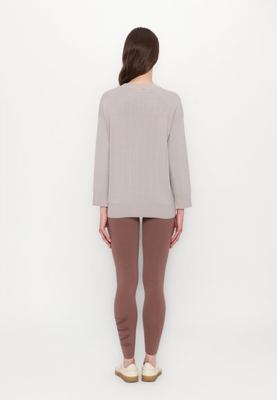 Pulover gri deschis cu țesătură în dungi, cu o croială lejeră, asortat cu colanți maro cu un model pe lateral. Modelul poartă încălțăminte clară.