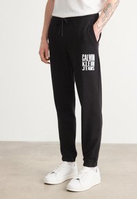 Melni sporta bikses no kokvilnas maisījuma, ar sašujamu jostasvietu un balto uzdrukāto "CALVIN KLEIN JEANS" logo kreisajā celiņā.