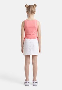 Παιδί με ξανθά μαλλιά σε δύο κότσους, φοράει ροζ αμάνικη μπλούζα, λευκή φούστα και λευκά αθλητικά παπούτσια, στέκεται με την πλάτη προς τα εμπρός σε λευκό φόντο.