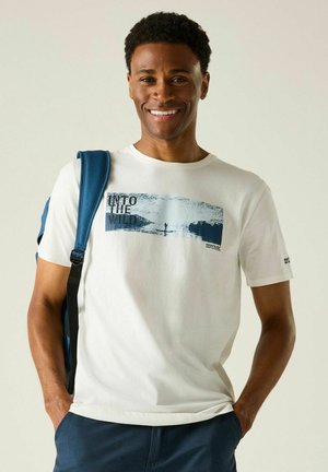Lächelnder Mann, der ein weißes "Into The Wild"-T-Shirt und einen blauen Rucksackriemen trägt, mit den Händen in den Taschen seiner blauen Hose, vor schlichtem Hintergrund stehend.