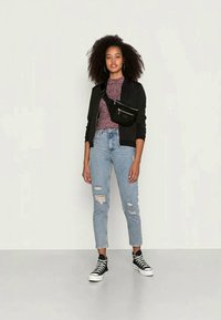 Jaqueta preta com fecho zip em cima de uma blusa texturada em bordeaux, combinada com jeans rasgados claros e tênis altos pretos. Bolsa de cinto preta com bolso frontal.