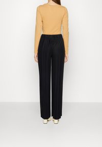 Kvinna som bär en senapsgul långärmad topp och högmidjade svarta plisserade vida byxor, stående mot en enfärgad vit bakgrund.