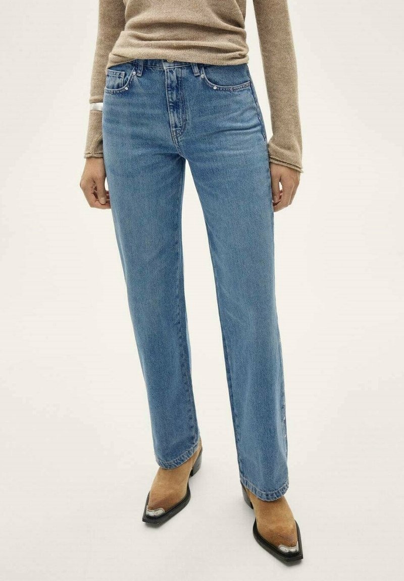 Femme portant un jean bleu clair à jambes droites, un haut beige à manches longues et des bottes pointues marron avec des accents argentés, debout sur un fond blanc.