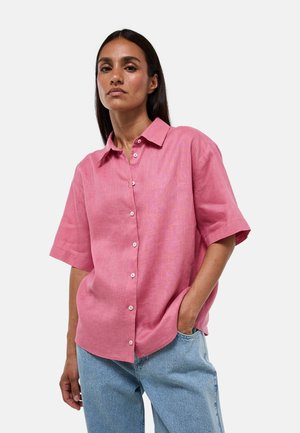 Femme aux longs cheveux foncés portant une chemise rose ample à manches courtes boutonnée et un jean bleu clair, une main dans la poche, sur fond uni.