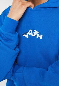 Niebieska bluza z kapturem wykonana z miękkiej tkaniny, z graficznym wzorem z napisem "zagubiona MŁODZIEŻ" w białym i czarnym kolorze, prezentująca swobodny styl.