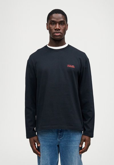 Sort langærmet t-shirt med hvid innerkrave, prydet med et rødt logo på venstre bryst. Båret med lyseblå denim jeans.