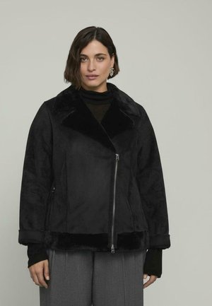 Mujer con cabello corto y oscuro, vestida con una chaqueta de piel sintética negra con cremallera, blusa negra, pantalones grises y pendientes en forma de hoja, sobre un fondo gris.