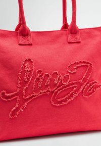 Roosa kangast kott, millel on suur, tekstuuriline logo tikand. Vastupidavad sangad ja ühtlane, veidi kare tekstuur kogu pinnal.