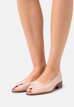Des chaussures ouvertes en cuir rose clair avec un petit talon bloc, présentant un design croisé à l'avant et une texture lisse.