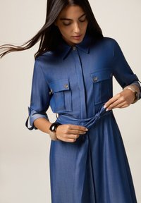 Una donna che sta allacciando una cintura su un vestito blu a maniche lunghe con tasche sul petto, indossando due braccialetti sul polso sinistro.