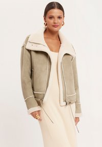 Kurtka z kołnierzem z sztucznego owczej wełny w miękkim oliwkowym kolorze z kontrastującymi białymi szwami, zamkiem błyskawicznym i regulowanym dołem.