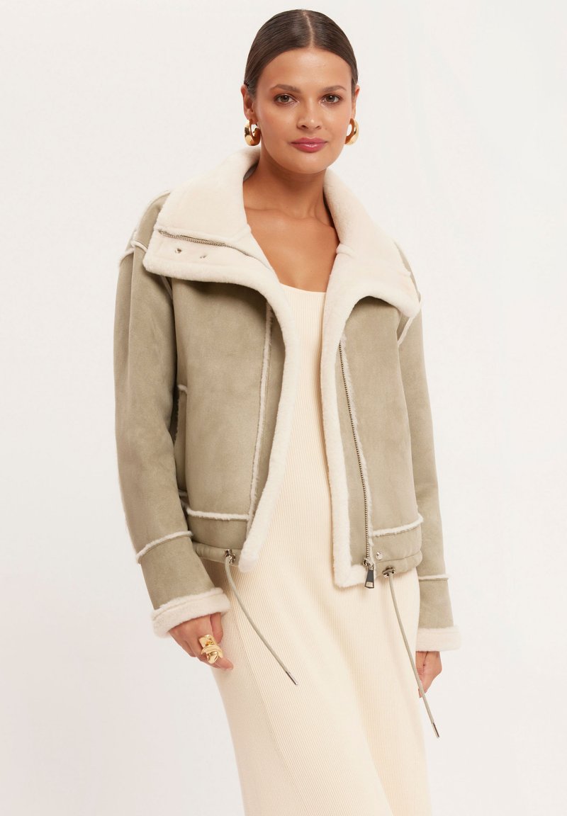 Kurtka z kołnierzem z sztucznego owczej wełny w miękkim oliwkowym kolorze z kontrastującymi białymi szwami, zamkiem błyskawicznym i regulowanym dołem.