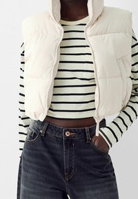Light beige puffer vest with hood over a black and white striped long-sleeve shirt, paired with high-waisted dark denim jeans.