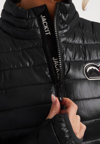 Gros plan sur une personne qui ferme une veste matelassée noire avec un col montant et un logo en patch sur la poitrine. Le curseur de la fermeture éclair indique "1."