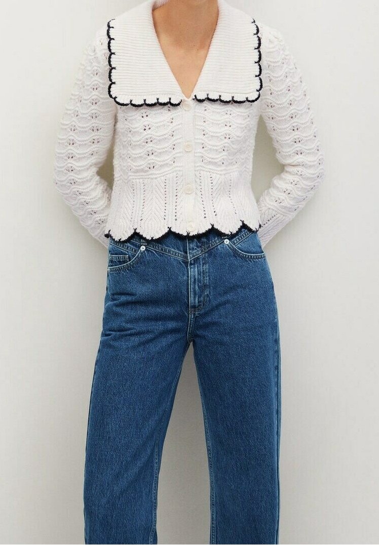 Cardigan blanc en tricot avec bords festonnés et grand col, associé à un jean bleu taille haute, mains dans les poches.
