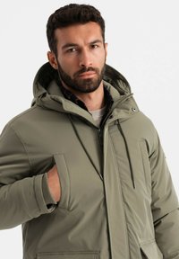 Olivengrüne isolierte Jacke mit Kapuze, ausgestattet mit Fronttaschen, einem zentralen Reißverschluss und verstellbaren Kordeln für einen sicheren Sitz.