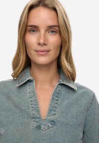 Světlá modrá džínová košile s výstřihem do V, strukturovaným límečkem a viditelnými švy. Textura látky je hladká s mírným leskem.