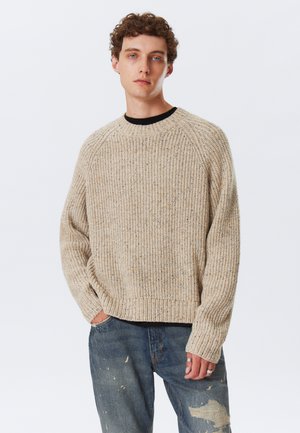 Beige knitted sweater with a ribbed texture, round neckline, and raglan sleeves, paired with distressed blue jeans.