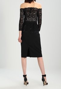Top en dentelle noire sans épaules avec des manches longues, taille peplum et jupe noire moulante, comportant une fente arrière et des talons hauts.