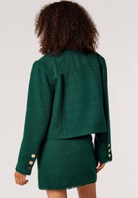 Grüner Tweedjacke mit einem verkürzten Design, fransigem Saum und langen Ärmeln. Goldene Knopfakzente an den Ärmelabschlüssen und strukturierten Oberflächen.