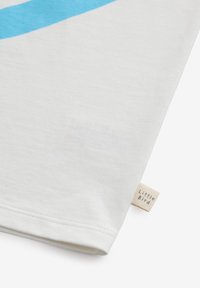 Vit bomulls T-shirt med ett stort blått diagonalt rand. Avslutad med en liten tygetikett märkt "Little Bird" vid nederkanten.