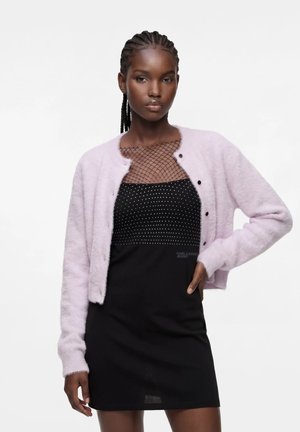 Femme aux cheveux tressés portant un cardigan duveteux violet clair par-dessus une robe noire avec un col en résille à pois, posant avec une main sur la hanche.