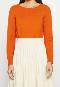 Orange-langärmliger Pullover aus weichem Material mit Rundhalsausschnitt, kombiniert mit einem plissierten cremefarbenen Rock mit breitem Bund.