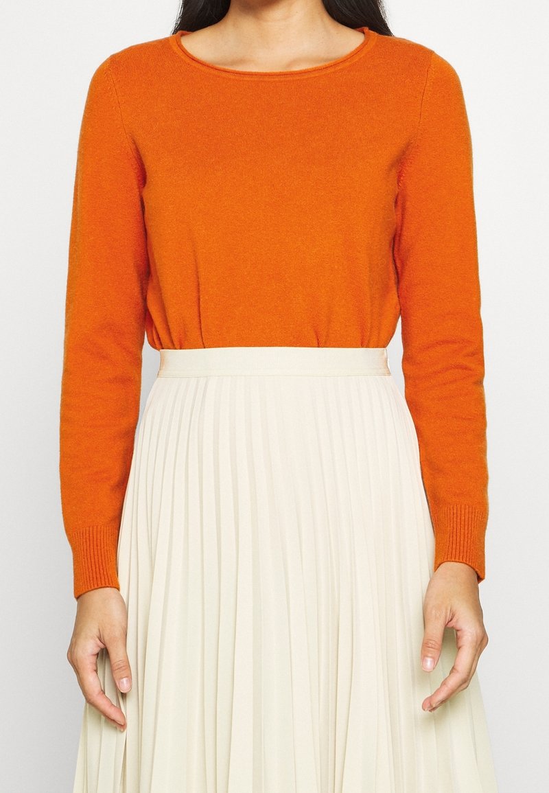 Orange-langärmliger Pullover aus weichem Material mit Rundhalsausschnitt, kombiniert mit einem plissierten cremefarbenen Rock mit breitem Bund.