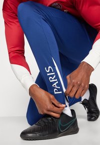 Pantalones deportivos azules con texto blanco "PARIS", combinados con una camiseta roja y blanca. El modelo ata los cordones de unas zapatillas deportivas negras.