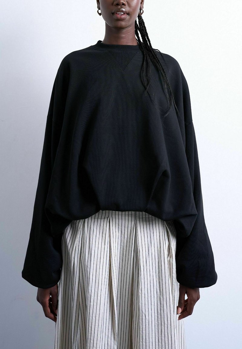 Mujer con cabello oscuro trenzado que lleva una sudadera negra oversize y una falda blanca de talle alto con finas rayas negras verticales.