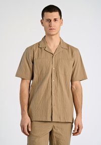 Camisa de manga corta en tejido texturizado marrón claro con cuello extendido, que presenta una parte delantera abotonada y dos bolsillos en el pecho.