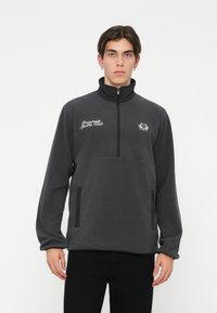Fleece pulover v temno sivi barvi z visokim ovratnikom, polovičnim zadrgo, dvema stranskima žepoma in vezenim logotipom "Originally Sport Club" na prsih.