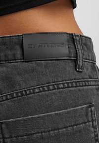 Jeans en denim noir avec une surface texturée, comportant une étiquette en cuir portant le logo "2Y STUDIOS" et des détails de couture visibles.