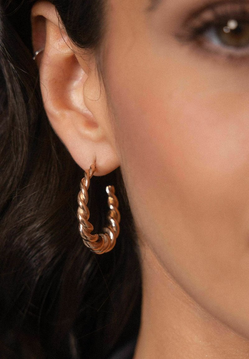 Kupferne, gedrehte Creole, die am Ohr getragen wird, mit einem spiralförmigen Design und glatter Textur, die ihren metallischen Glanz und ihre Form betont.