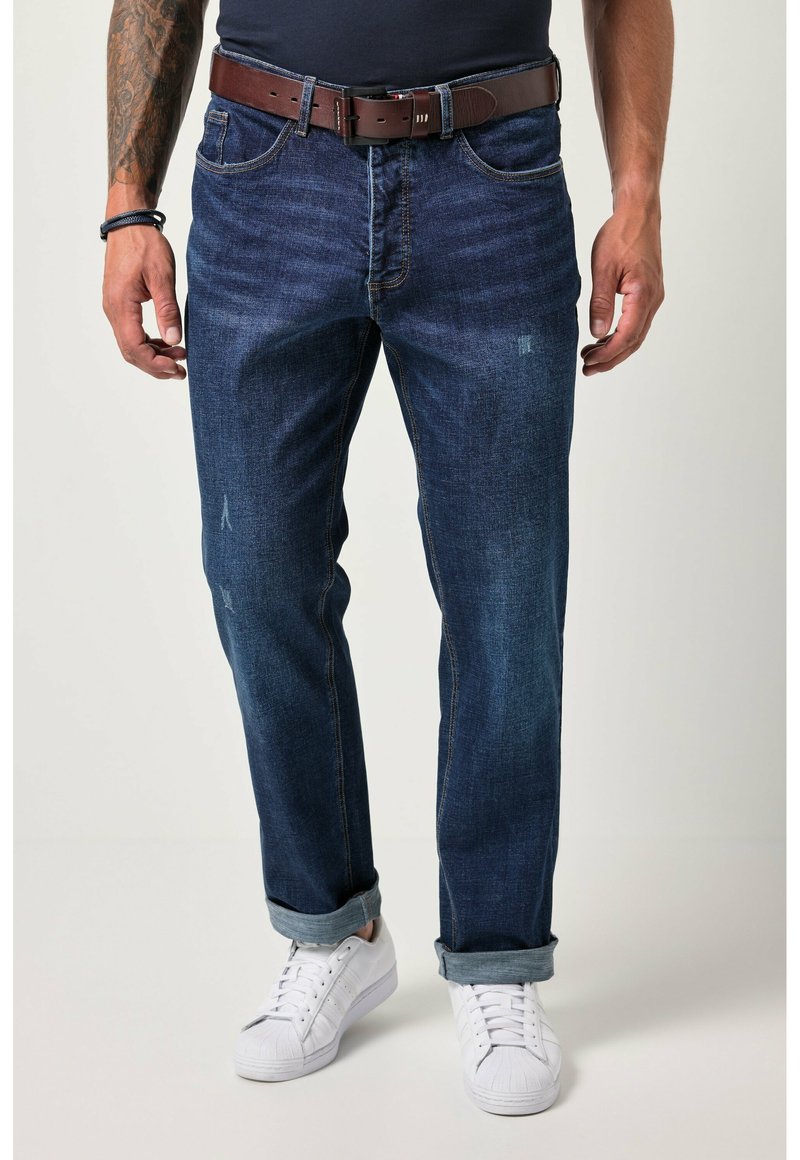 Hombre que lleva unos jeans oscuros de corte recto, cinturón de cuero marrón, zapatillas blancas y una camisa negra, de pie con los brazos relajados a los lados.