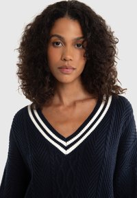Maglione navy con un motivo testurizzato, dotato di scollo a V e accenti a strisce bianche sul colletto. Il materiale sembra lavorato a maglia e spesso.