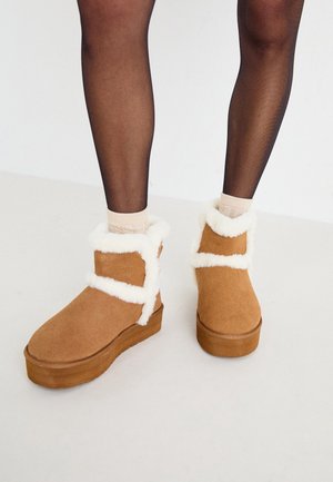Bottes cheville en daim beige avec doublure en fausse fourrure blanche et accents de lanières ; semelle plate avec une légère épaisseur. Jambes en collants noirs transparents.