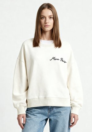 Ung kvinde med glat brunt hår iført en stor hvid sweatshirt med teksten "Mira Paris" kombineret med lyseblå jeans, stående neutralt.